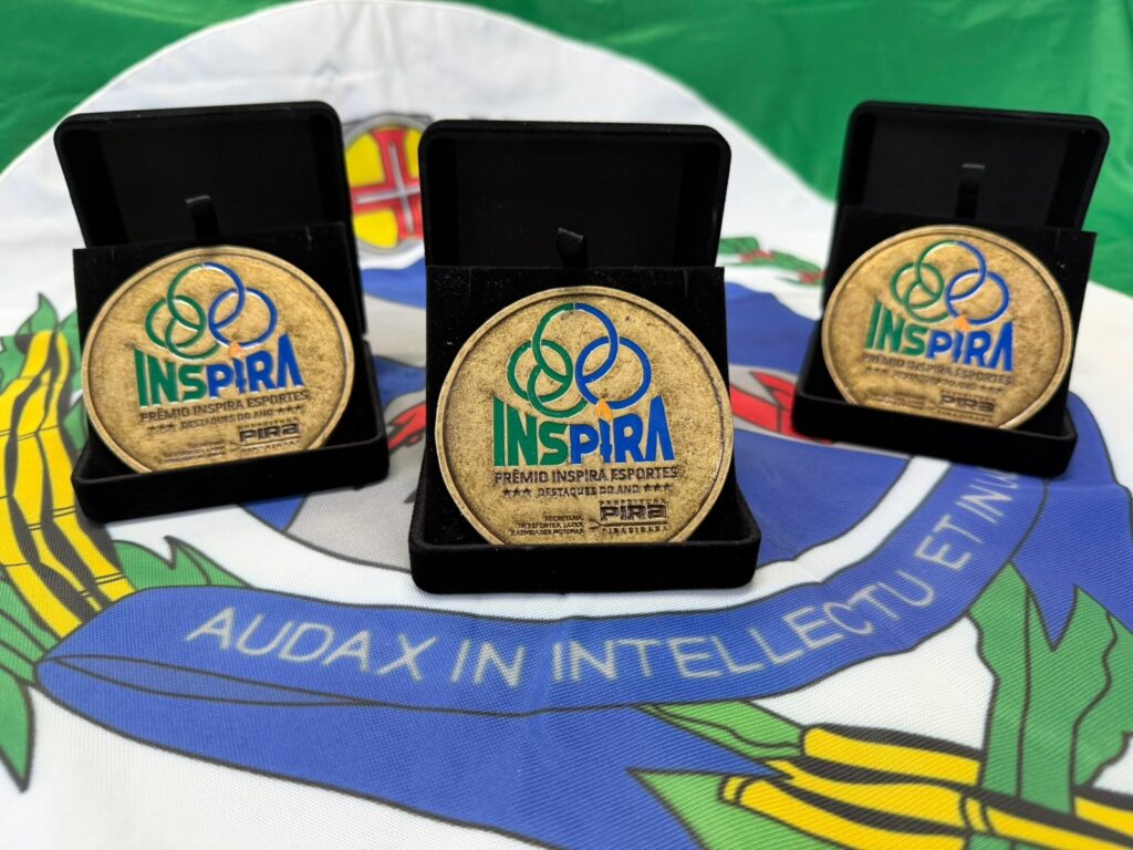 A imagem exibe três medalhas douradas, cada uma em uma caixa preta aberta, dispostas sobre uma bandeira. As medalhas apresentam o logo "INSPIRA" em verde e azul, com a inscrição "PRÊMIO INSPIRA ESPORTES" e "DESTAQUES DO ANO". Abaixo, lê-se "PREFEITURA PIRA", indicando a cidade de Piracicaba. A bandeira sobre a qual as medalhas repousam é a do município de Piracicaba, com suas cores e o lema "AUDAX IN INTELLECTU ET IN RE". A fotografia tem um foco suave, destacando o brilho das medalhas e os detalhes da bandeira.