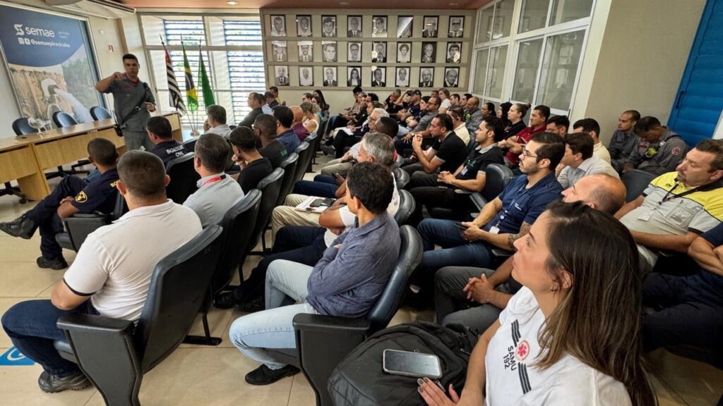 A imagem mostra uma sala de treinamento com um homem uniformizado, presumivelmente um instrutor, em pé e gesticulando para uma audiência sentada em cadeiras. O instrutor veste um uniforme cinza com detalhes em vermelho e preto, e parece estar explicando algo. A audiência é composta por homens e mulheres, muitos deles vestindo uniformes que sugerem serem bombeiros civis ou profissionais de serviços de emergência. As cadeiras estão dispostas em fileiras, voltadas para o instrutor. Ao fundo, há uma parede com várias fotografias emolduradas, possivelmente de figuras importantes ou históricas. Há também bandeiras e um grande painel com