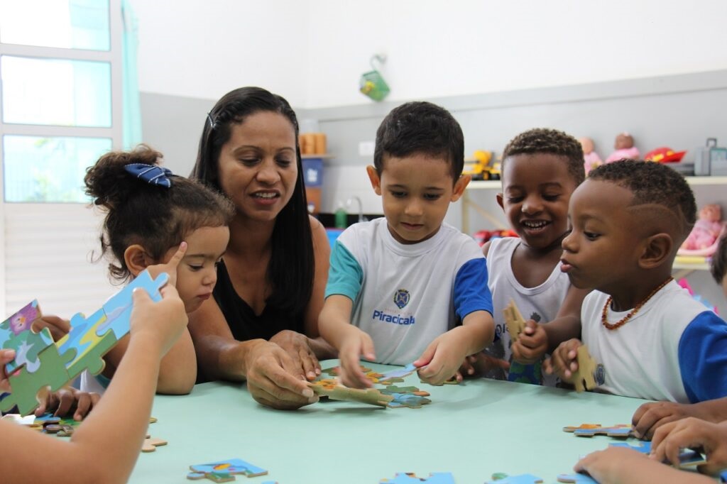 A imagem mostra um grupo de crianças com uma professora em uma sala de aula, todas reunidas em torno de uma mesa para montar um quebra-cabeça. A professora, uma mulher morena com cabelo preto preso, está sorrindo e olhando para as crianças. Há quatro crianças visíveis, todas concentradas no quebra-cabeça. Uma menina com um laço azul no cabelo está segurando uma peça do quebra-cabeça e apontando para ela. Um menino com uma camiseta branca com detalhes azuis e verdes, onde se lê "Piracicaba", está manipulando as peças do quebra-cabeça. Ao lado dele, outro menino com uma camiseta branca e azul também está engajado na atividade, sorrindo. Um terceiro menino, com uma camiseta branca, está olhando para o quebra-cabeça com atenção. A mesa é verde e várias peças do quebra-cabeça estão espalhadas sobre ela. Ao fundo, pode-se ver uma janela com cortinas azuis claras e prateleiras com brinquedos e materiais escolares. A atmosfera geral da imagem é de aprendizado e diversão em um ambiente escolar.