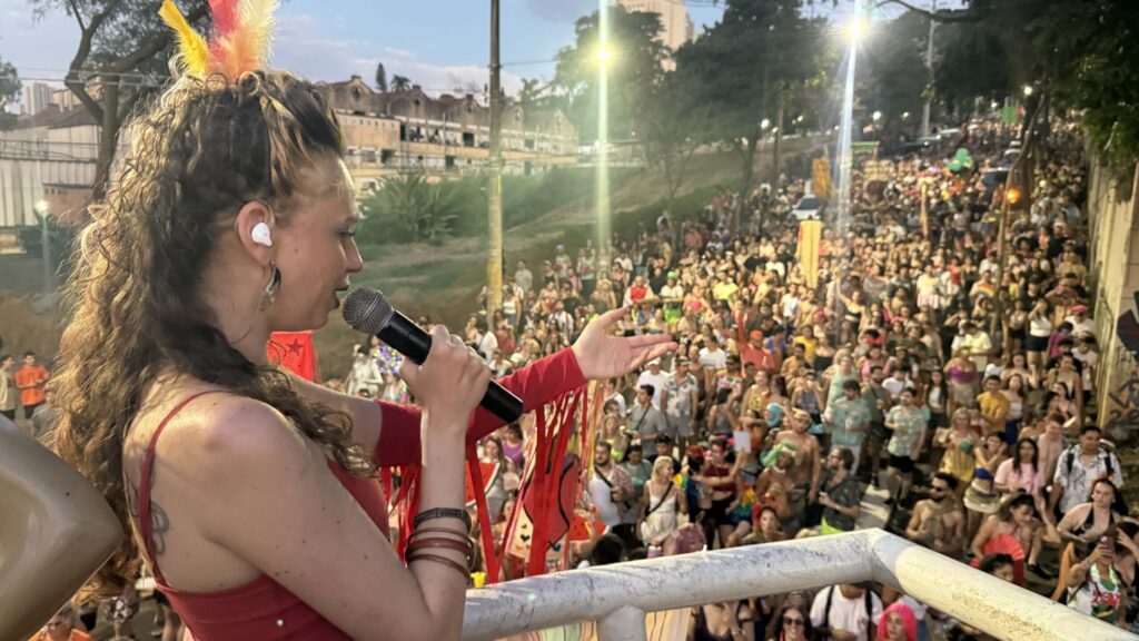 A imagem mostra uma cantora em primeiro plano, com um microfone na mão e um arranjo de penas coloridas em seu cabelo. Ela está vestindo uma roupa vermelha com franjas. Atrás dela, uma multidão de pessoas celebra em um evento ao ar livre, possivelmente um carnaval ou festival. O ambiente parece ser urbano, com prédios ao fundo e iluminação artificial. A atmosfera é vibrante e festiva, com muitas pessoas vestindo roupas coloridas e expressando alegria.