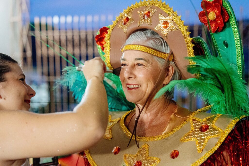 A imagem mostra uma mulher mais velha, com cabelos grisalhos, sorrindo enquanto usa um traje de carnaval elaborado. Ela usa um chapéu grande e decorado com estrelas douradas e detalhes vermelhos e dourados brilhantes. O traje em si é dourado, com bordas douradas e estrelas semelhantes, adornado com pedras vermelhas e laranjas. Penas verdes vibrantes emanam de suas costas e adornam o chapéu, adicionando um toque de cor e exuberância. Outra mulher, vista de perfil no lado esquerdo da imagem, está ajustando o traje da mulher mais velha, tocando nas penas verdes. A iluminação sugere que a cena pode estar ocorrendo em um ambiente interno, possivelmente um camarim ou um local de preparação para um evento festivo. O fundo é desfocado, com luzes e formas que indicam um ambiente movimentado. A expressão feliz da mulher mais velha sugere que ela está gostando do momento e está animada para o evento.