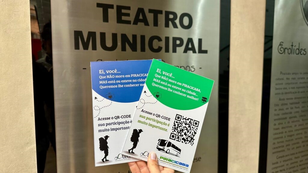 A imagem mostra duas pequenas brochuras, uma azul e outra verde, sendo seguradas por uma mão. Ambas as brochuras têm o mesmo texto e design, com exceção da cor. O texto principal diz: "Ei, você... Que NÃO mora em PIRACICABA, MAS está ou esteve na cidade... Queremos lhe conhecer melhor!". Abaixo deste texto, há uma instrução: "Acesse o QR-CODE sua participação é muito importante." Um código QR está impresso em ambas as brochuras, e abaixo dele há uma ilustração de um ônibus e o logotipo "PIRACICABA". As brochuras estão posicionadas em frente a um letreiro grande que diz "TEATRO MUNICIPAL". Ao lado direito do letreiro, há um outro cartaz com o título "Grotides" e texto em português, embora este esteja parcialmente obscurecido e fora de foco. O fundo geral sugere que as brochuras estão sendo oferecidas ou distribuídas em um evento ou local público, possivelmente no próprio Teatro Municipal.