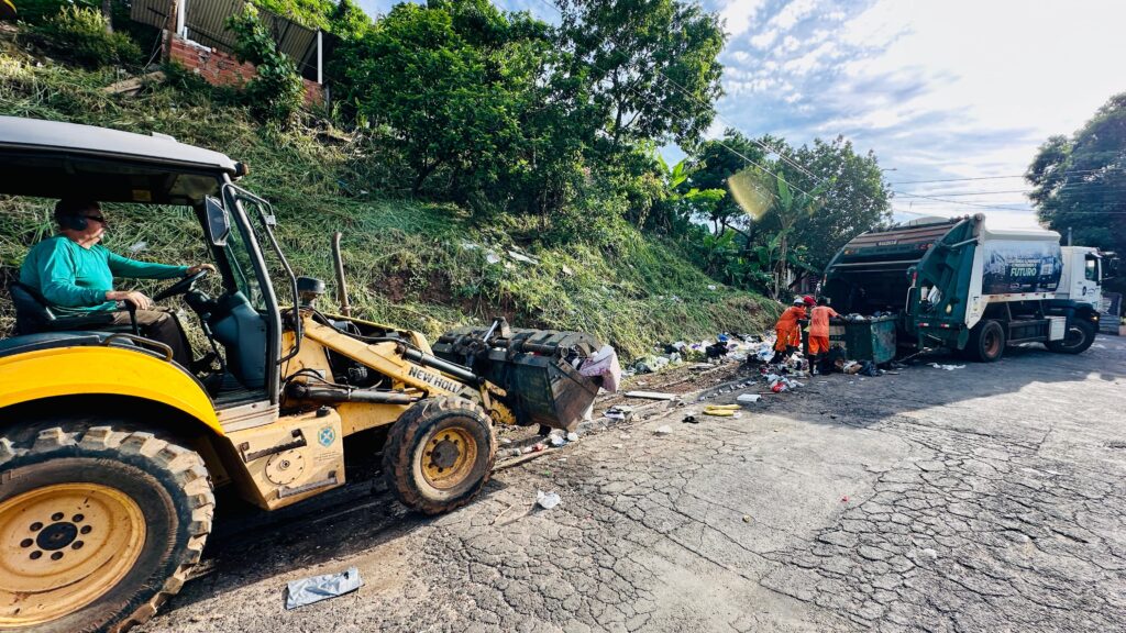 A imagem mostra uma operação de coleta de lixo às margens de uma estrada, envolvendo diferentes equipamentos e trabalhadores. À esquerda, um trator carregador amarelo movimenta resíduos acumulados, enquanto à direita dois garis, vestidos com uniformes laranja, colocam o lixo em um caminhão verde e branco. O cenário inclui um trecho de asfalto danificado, restos espalhados pelo chão e, ao fundo, uma encosta com árvores e construções.