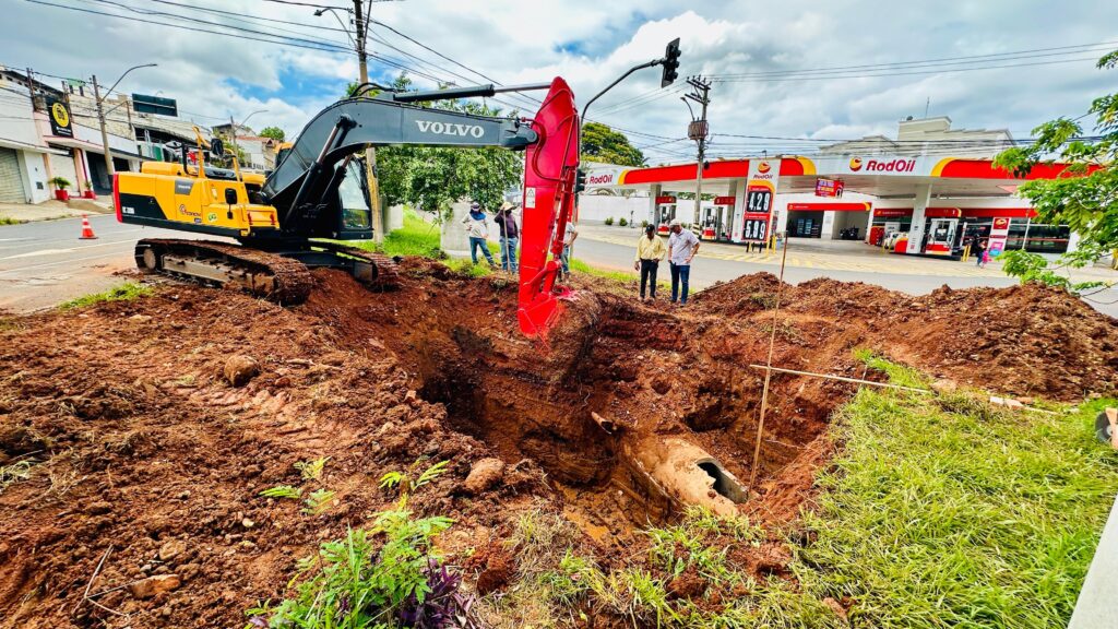 A imagem mostra uma escavadeira Volvo amarela e preta em um canteiro de obras, com uma grande vala aberta. A escavadeira está posicionada de forma que sua lança e balde vermelhos estejam sobre a vala, possivelmente em processo de escavação ou remoção de terra. Várias pessoas estão reunidas perto da vala, observando o trabalho. Ao fundo, é possível ver um posto de gasolina RodOil com suas bombas e a fachada do estabelecimento. Há também alguns edifícios e postes de iluminação pública, indicando que a cena se passa em uma área urbana ou suburbana. O céu está parcialmente nublado, sugerindo um dia com clima variável. A terra ao redor da vala é de cor marrom-avermelhada, típica de solo argiloso, e há vegetação verde crescendo nas bordas da vala e no primeiro plano. Um cone de trânsito laranja está visível na estrada, próximo à escavadeira, indicando a sinalização de obra.