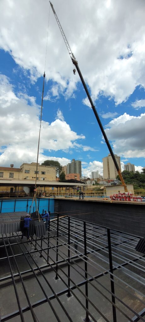 A fotografia mostra uma cena de construção, com um guindaste imponente em destaque contra um céu azul vibrante pontuado por nuvens brancas fofas. O braço do guindaste se estende diagonalmente pela imagem, com seu gancho pendurado, pronto para levantar ou abaixar cargas. Na parte inferior da imagem, um grande tanque ou reservatório está em construção. Trabalhadores com capacetes azuis podem ser vistos manuseando componentes escuros e retangulares, possivelmente painéis ou blocos, que estão sendo baixados para o interior do tanque. Uma estrutura metálica complexa, composta por barras e suportes, já está montada no fundo do reservatório, indicando o progresso da obra. Ao fundo, edifícios de vários andares compõem a paisagem urbana, sugerindo que a construção está ocorrendo em uma área desenvolvida. A combinação do céu dramático, a maquinaria pesada e a atividade humana criam uma sensação de empreendimento e progresso.