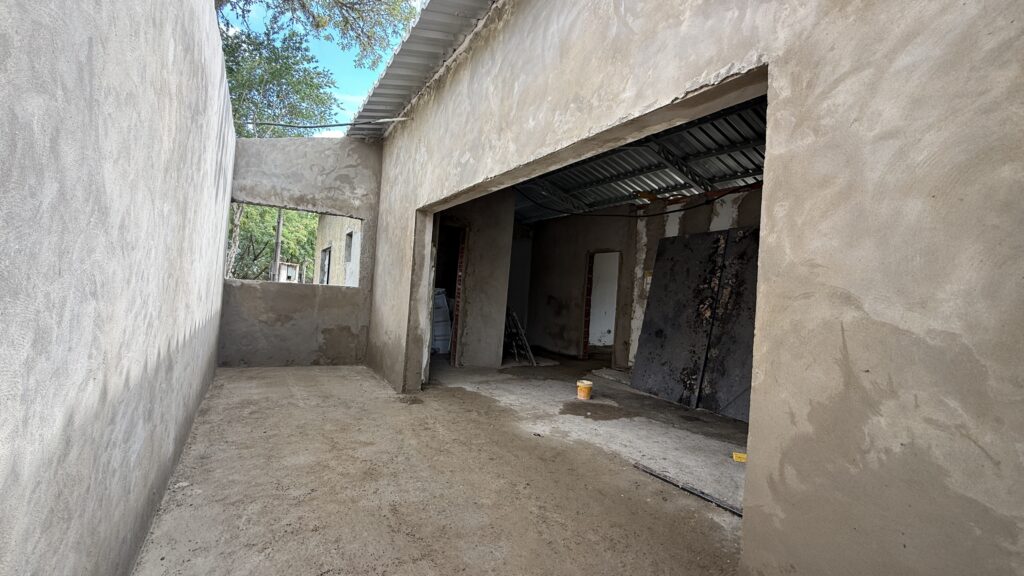 A imagem mostra o interior de uma construção em andamento. As paredes estão rebocadas, mas ainda não pintadas, e o chão é de cimento bruto. Há uma abertura ampla que dá para outra área da construção, onde se pode ver o telhado de metal aparente e algumas portas. Uma das paredes internas possui uma janela retangular que permite a visão do exterior, onde árvores e o céu azul são visíveis. Há alguns materiais de construção espalhados pelo chão, incluindo um balde amarelo e chapas escuras apoiadas em uma parede. A iluminação sugere que é dia, com luz natural entrando pelas aberturas. O ambiente geral é de uma obra em fase inicial de acabamento.