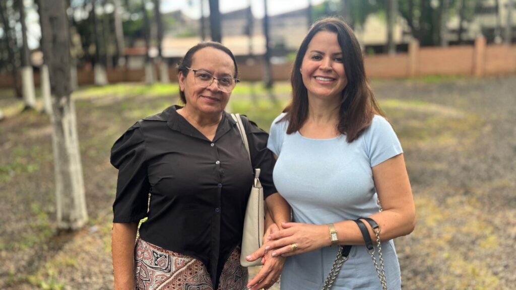 A imagem mostra duas mulheres sorrindo para a câmera, em pé ao ar livre. A mulher à esquerda, mais velha, usa óculos, uma camisa preta de botões com mangas bufantes e uma saia estampada. Ela carrega uma bolsa bege sobre o ombro. A mulher à direita, mais jovem, usa uma blusa azul clara de gola redonda e calças ou shorts azuis claros. Ela usa um relógio no pulso esquerdo e uma bolsa com alça de corrente preta. Ambas as mulheres parecem estar de mãos dadas ou com os braços entrelaçados, transmitindo uma sensação de proximidade e afeto. O fundo é desfocado, mostrando árvores e um caminho de cascalho, sugerindo um ambiente natural ou um parque. A iluminação é suave, indicando um dia nublado ou luz indireta.