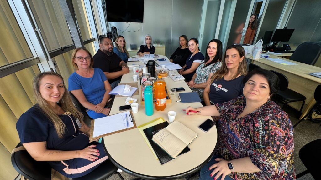 A imagem mostra um grupo de pessoas sentadas ao redor de uma mesa em uma sala de reuniões. Há nove pessoas visíveis na imagem, a maioria mulheres, com um homem sentado à esquerda. A mesa está coberta com papéis, cadernos, canetas, copos, uma garrafa de refrigerante e uma garrafa térmica. Há também um bolo em uma bandeja. A iluminação é natural, vindo de grandes janelas ao fundo, que parecem ter um design arquitetônico interessante com barras verticais. Em segundo plano, há um escritório com mesas, computadores e cadeiras. Uma mulher está em pé em uma porta de vidro ao fundo, olhando para dentro da sala. A atmosfera parece ser de uma reunião de trabalho ou um encontro informal.