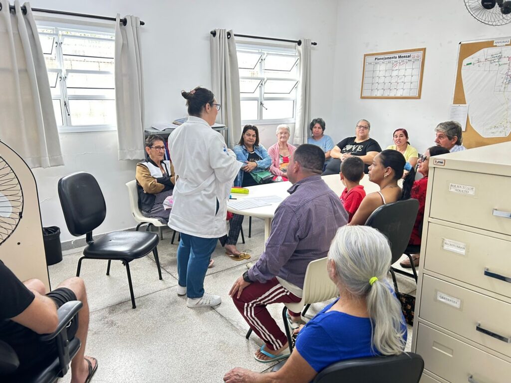 A imagem mostra um ambiente interno que parece ser uma clínica ou centro de saúde comunitário. Há um grupo de pessoas sentadas em cadeiras ao redor de uma mesa branca oval e também ao longo das paredes. A maioria são adultos, com algumas pessoas mais velhas e uma criança pequena. Uma mulher vestida com jaleco branco, calça jeans e tênis brancos está em pé, de costas para a câmera, aparentemente falando ou orientando o grupo. O ambiente é claro, com paredes brancas e duas janelas com cortinas claras. No fundo, há um armário metálico bege com gavetas identificadas como "FOLHA ROSTO ADULTO", "RECEITUÁRIOS" e "GESTANTES". Também há um quadro na parede com um planejamento mensal e um mapa com o nome "ANHUMAS". A iluminação é fluorescente, conferindo ao local uma aparência limpa e organizada. A cena sugere uma reunião ou palestra educativa, possivelmente relacionada à saúde ou a algum programa comunitário, onde a profissional de saúde está passando informações para os presentes.