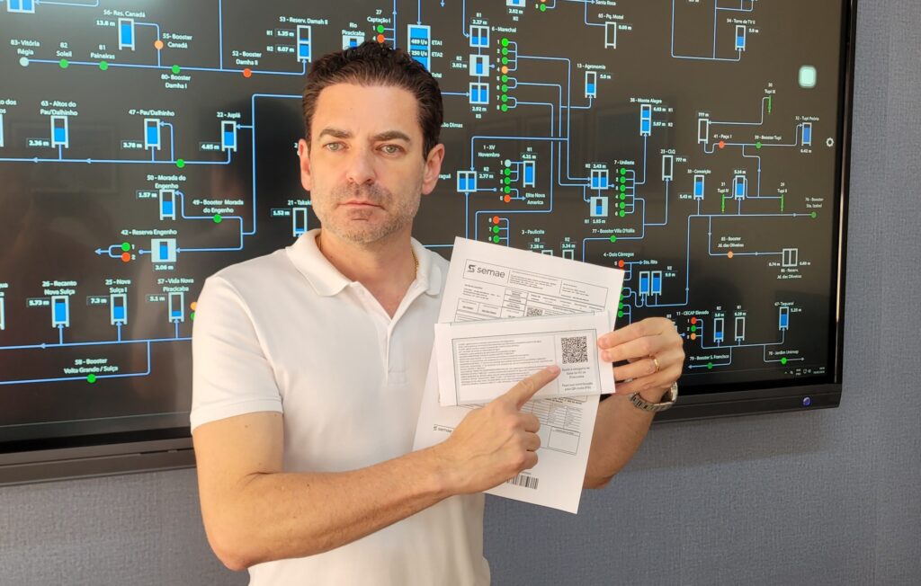 O homem na foto está em frente a uma grande tela de monitor que exibe um mapa do sistema de abastecimento de água, com vários reservatórios e estações de bombeamento identificados por números e nomes. Ele está vestindo uma camisa polo branca e segura alguns papéis nas mãos, apontando para um deles com o dedo indicador. Os papéis parecem ser contas ou documentos informativos, com um código QR visível em um deles. A tela ao fundo mostra um diagrama complexo, indicando a interconexão e o status de diferentes pontos do sistema de água