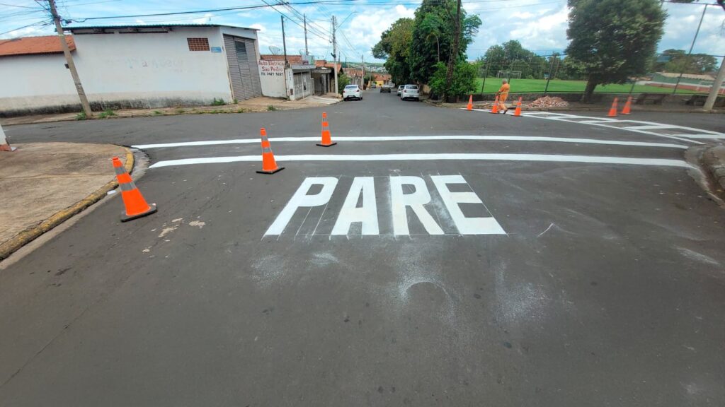 A imagem mostra um cruzamento de rua com sinalização recém-pintada. No centro da imagem, a palavra "PARE" está pintada em letras brancas grandes no asfalto escuro, indicando a necessidade de parar. Várias faixas brancas foram pintadas no asfalto, formando uma faixa de pedestres e delimitando o cruzamento. Cones de trânsito laranja estão posicionados estrategicamente ao redor da área para direcionar o tráfego e alertar os motoristas sobre a nova sinalização. Ao fundo, é possível ver carros estacionados e em movimento, edifícios com fachadas claras e um poste de eletricidade. Há também uma área gramada com uma quadra esportiva e uma pessoa vestindo um uniforme laranja, possivelmente um trabalhador da construção ou manutenção, próximo a uma pilha de tijolos. O céu está parcialmente nublado, com algumas nuvens visíveis.