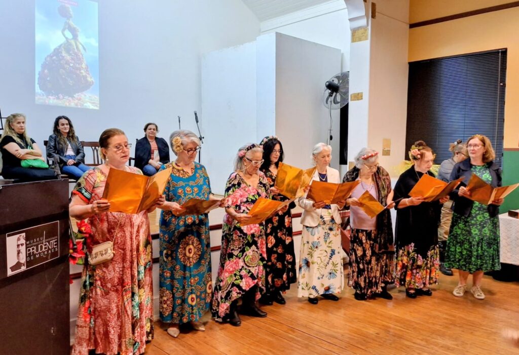 O grupo de mulheres se apresenta em um palco, segurando partituras douradas. Elas estão vestidas com trajes vibrantes e floridos, cada uma com um estilo único. Algumas usam flores nos cabelos, adicionando um toque festivo à apresentação. Ao fundo, um telão exibe uma imagem artística, possivelmente relacionada ao tema da apresentação. Há também cadeiras onde algumas pessoas estão sentadas, assistindo à performance. À esquerda, um púlpito com o logo do "Museu Prudente de Moraes" indica o local do evento. A iluminação e a disposição dos elementos criam uma atmosfera de evento cultural e artístico.