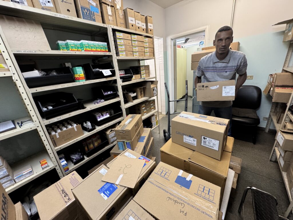 A imagem mostra um homem segurando uma caixa em um armazém ou depósito, com prateleiras cheias de caixas e suprimentos médicos ao fundo. As prateleiras estão organizadas com vários produtos, incluindo caixas de medicamentos e suprimentos de laboratório. O homem está em pé, de frente para a câmera, e parece estar em processo de movimentação de caixas. Há um carrinho de mão próximo, sugerindo que o trabalho envolve logística e organização de estoque. O ambiente parece ser um local de armazenamento de produtos farmacêuticos ou de saúde, dada a natureza dos itens visíveis nas prateleiras.