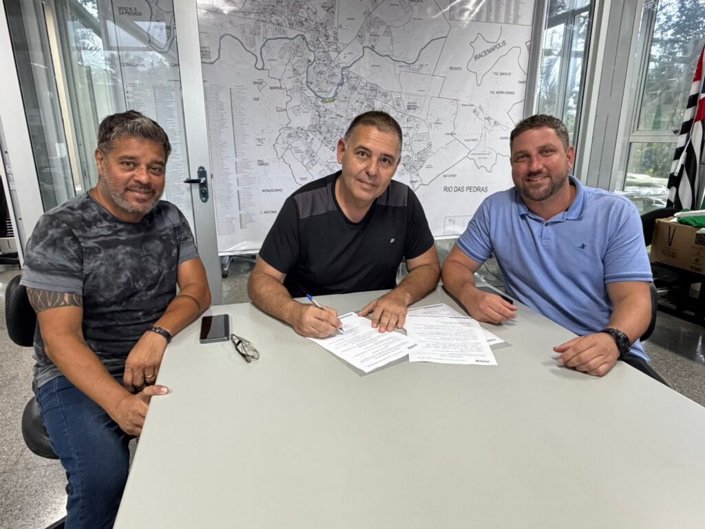 A imagem mostra três homens sentados em uma mesa, com um mapa ao fundo. O homem da esquerda, com uma camiseta tie-dye cinza e jeans, sorri para a câmera. O homem do meio, vestindo uma camiseta preta, está assinando um documento. O homem da direita, com uma camiseta azul clara, sorri para a câmera. Há também um celular, óculos e documentos sobre a mesa. Ao fundo, um mapa detalhado da região, com nomes de cidades e rios, e uma bandeira do estado de São Paulo.