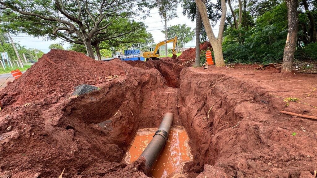 A imagem mostra um canteiro de obras com uma vala profunda escavada no solo vermelho. Um cano grande está parcialmente submerso em água lamacenta dentro da vala. Montanhas de terra vermelha, resultado da escavação, cercam a vala. Ao fundo, um escavador amarelo está posicionado perto de um grande tanque azul com a palavra "DEPINI" escrita. Uma escada está apoiada em uma das bordas da vala. Há também cones de trânsito laranja e alguns materiais de construção espalhados pela área. Árvores e vegetação exuberante criam um cenário natural ao redor da obra. O céu está nublado, sugerindo um dia úmido.