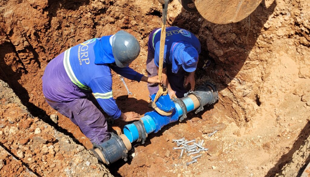 A imagem mostra dois trabalhadores em um fosso cavado no solo, realizando a instalação de uma tubulação. Ambos vestem uniformes azuis e um deles usa um capacete cinza. A tubulação em questão é composta por seções de PVC azul e conexões pretas, com uma peça central azul que parece ser uma válvula ou registro. Uma das peças da tubulação está sendo sustentada por uma cinta amarela, indicando que está sendo posicionada ou ajustada. Ao lado da tubulação, há um monte de parafusos metálicos, provavelmente destinados à fixação das conexões. O solo ao redor é de cor marrom avermelhada, com algumas pedras e areia visíveis. A iluminação sugere que a imagem foi tirada durante o dia, com sombras projetadas no fosso.