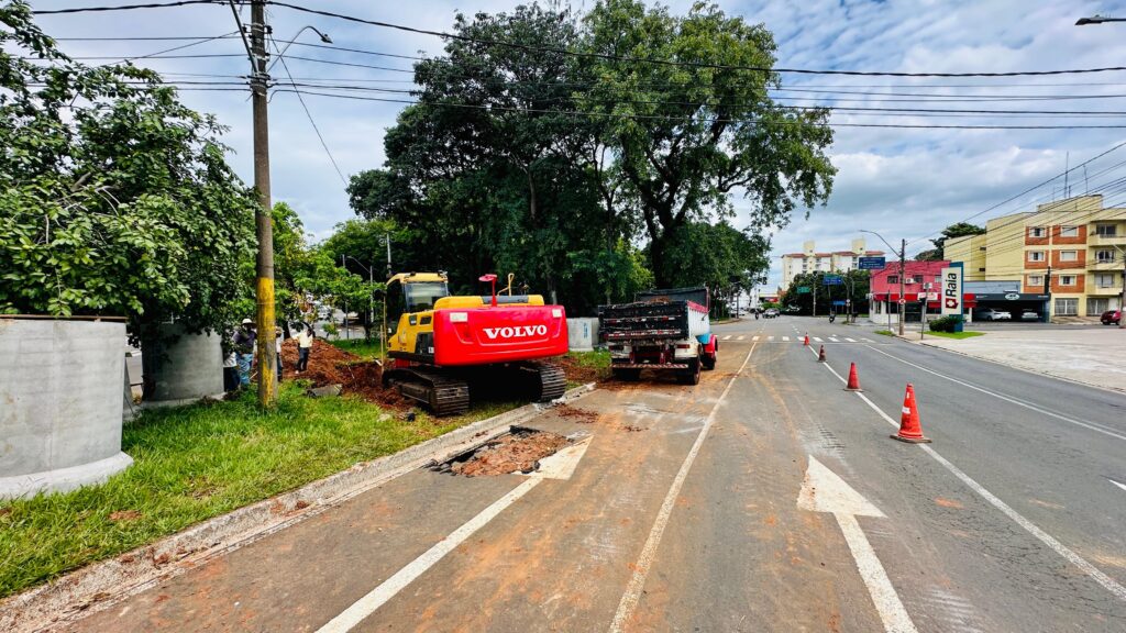 A imagem mostra uma cena de obras em uma rua urbana. No primeiro plano, um escavador Volvo amarelo e vermelho está posicionado ao lado de uma vala recém-aberta na pista. Ao lado do escavador, um caminhão de caçamba está estacionado, pronto para transportar terra ou material. Na lateral da rua, há uma área gramada com duas grandes estruturas cilíndricas de concreto, possivelmente parte de um sistema de saneamento ou drenagem. Várias pessoas, vestindo roupas de trabalho e capacetes, estão supervisionando a obra. O fundo da imagem revela a paisagem urbana com edifícios, árvores frondosas e postes de iluminação com fiação aérea. O céu está nublado, mas a luz do dia sugere que o trabalho está em andamento. A sinalização de trânsito, incluindo cones laranja, indica que a área está em obras e o tráfego pode ser afetado. Setas brancas pintadas na pista indicam a direção do tráfego.