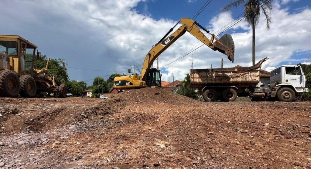 A imagem mostra um cenário de construção em uma área externa, sob um céu azul com nuvens brancas. Há duas máquinas pesadas em operação: uma escavadeira amarela na frente, com seu braço estendido e a concha sobre uma pilha de terra, e um nivelador (grader) amarelo atrás, com suas rodas e lâmina visíveis. Ao fundo, observa-se uma construção baixa, com telhado de metal e paredes claras, onde se destacam uma janela com grades brancas e uma antena parabólica no telhado. Há também vegetação verde e árvores ao redor, além de alguns postes e fios elétricos. A terra na frente das máquinas parece ter sido remexida, indicando trabalho em andamento.