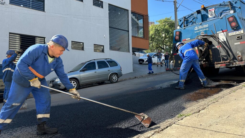 A imagem mostra trabalhadores em um dia ensolarado, realizando obras de pavimentação em uma rua urbana. * No primeiro plano, um trabalhador de macacão azul e capacete, com luvas e botas de segurança, está espalhando asfalto com uma pá. * Ao fundo, outro trabalhador está posicionado perto de um caminhão de asfalto azul, que parece estar descarregando o material. O caminhão possui a identificação "Mólise Serviços e Construções Ltda." e a placa "OOC8C54". * Há carros estacionados e em movimento na rua, e edifícios residenciais ao lado da via. * A cena retrata o trabalho de manutenção e construção de vias públicas.