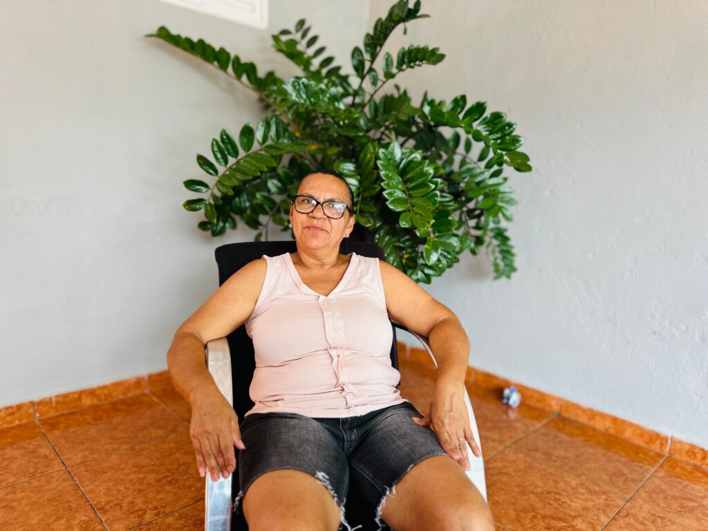 A mulher senta-se numa cadeira preta com braços de metal, vestida com uma regata rosa pálido e calções de ganga cinzentos. Ela usa óculos pretos e tem o cabelo escuro preso para trás. O seu olhar está dirigido para a câmara com um leve sorriso. Atrás dela, um grande arbusto de folhas verdes compridas e brilhantes cresce contra uma parede de cor creme. O chão à frente dela é coberto por azulejos cor de terracota dispostos em padrão de espinha. No canto inferior direito, um pequeno objeto azul e branco está no chão. A iluminação geral sugere um ambiente exterior ou um espaço interior bem iluminado com luz natural.