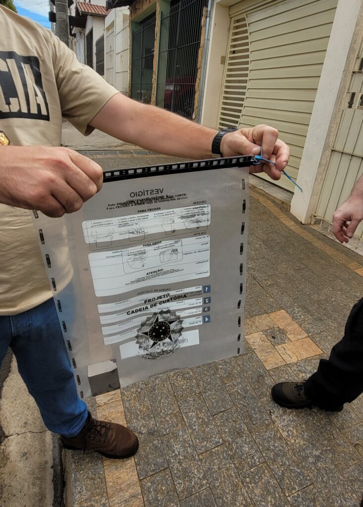 A imagem mostra um indivíduo, vestindo uma camiseta com a palavra "POLICIA" estampada, segurando um envelope transparente com instruções de fechamento e abertura. O envelope faz parte de um "PROJETO CADEIA DE CUSTÓDIA" e possui o brasão da República Federativa do Brasil. O indivíduo está amarrando o envelope com um lacre azul. Ao fundo, observa-se uma rua com casas. Outra pessoa, com calças pretas e sapatos escuros, está parcialmente visível na lateral direita da imagem.