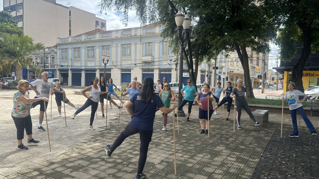 A imagem mostra um grupo de pessoas, a maioria idosos, participando de uma aula de exercícios ao ar livre em uma praça. Eles estão em pé, usando bastões de madeira para se equilibrar enquanto realizam movimentos que parecem alongamentos ou exercícios de mobilidade. O ambiente é urbano, com edifícios ao fundo que apresentam arquitetura clássica, alguns com detalhes em azul claro. Há árvores frondosas, postes de iluminação e calçamento de pedras no chão, criando uma atmosfera de parque ou praça pública. As pessoas vestem roupas casuais e esportivas e parecem engajadas na atividade, com expressões de concentração e esforço. Uma instrutora, de costas para a câmera, parece estar guiando o grupo.