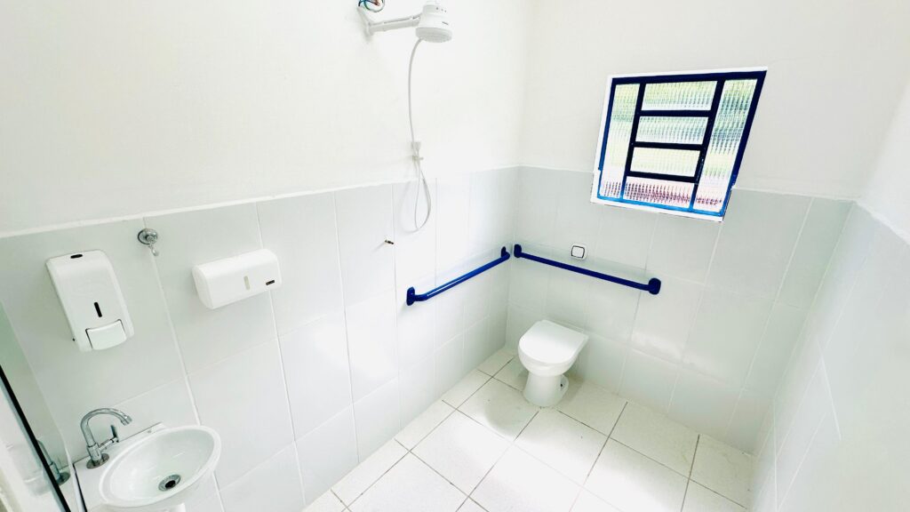 [16:10, 21/01/2026] IA: Analisando a imagem, me dê um momento... 🕵️
[16:10, 21/01/2026] IA: A imagem mostra um banheiro acessível, com paredes e piso revestidos de azulejos brancos. Há um vaso sanitário com barras de apoio azuis instaladas nas paredes ao redor, para auxiliar pessoas com mobilidade reduzida.No lado esquerdo, há uma pequena pia com torneira cromada, um dispensador de sabão líquido e um porta-papel. Acima da pia, um gancho para pendurar algo.No canto superior esquerdo, uma ducha elétrica está instalada na parede, com seu chuveiro posicionado para uso.No canto superior direito, uma janela com grades e vidro texturizado permite a entrada de luz natural.O ambiente é limpo e funcional, projetado para atender às necessidades de acessibilidade.