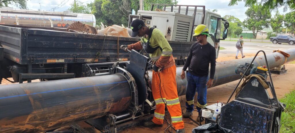 A imagem mostra dois trabalhadores em um canteiro de obras, aparentemente realizando a instalação de um grande tubo de plástico preto com uma linha azul. Um trabalhador, vestindo um chapéu verde, um colete amarelo e calças laranja de alta visibilidade, está operando uma máquina que parece estar conectada ao tubo. Ele usa luvas e está focado em sua tarefa. O outro trabalhador, usando um chapéu amarelo e uma camiseta preta de manga comprida com calças azuis, está observando o processo. Ao fundo, um caminhão basculante com uma carga de metal e um tanque de combustível visível. Há também um gerador grande no caminhão. A área ao redor parece ser um local de construção, com terra e grama visíveis. Ao longe, uma estrada com tráfego e árvores sugerem um ambiente urbano ou suburbano. O dia está nublado, mas claro. Um terceiro indivíduo, vestindo roupas casuais e carregando uma mochila, caminha pela área em segundo plano. Há equipamentos de soldagem e outros materiais de construção espalhados pelo chão perto dos trabalhadores.