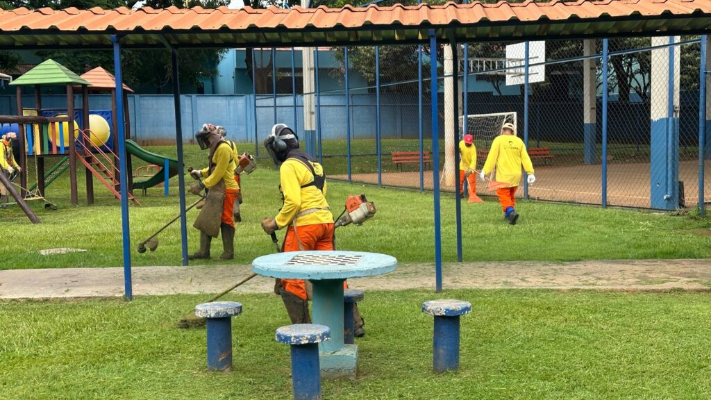 A imagem mostra um grupo de trabalhadores em um parque ou área de lazer, todos vestidos com uniformes amarelos e calças laranjas. Alguns deles estão usando cortadores de grama, enquanto outros estão varrendo o chão. No fundo, há um playground com um escorregador e balanços, além de uma quadra esportiva com uma cesta de basquete e uma trave de futebol. Há também uma mesa de piquenique com bancos ao redor, todos com uma pintura azul desbotada. A área está gramada e parece estar bem cuidada, com os trabalhadores realizando tarefas de manutenção. O céu está nublado, o que sugere um dia mais fresco.