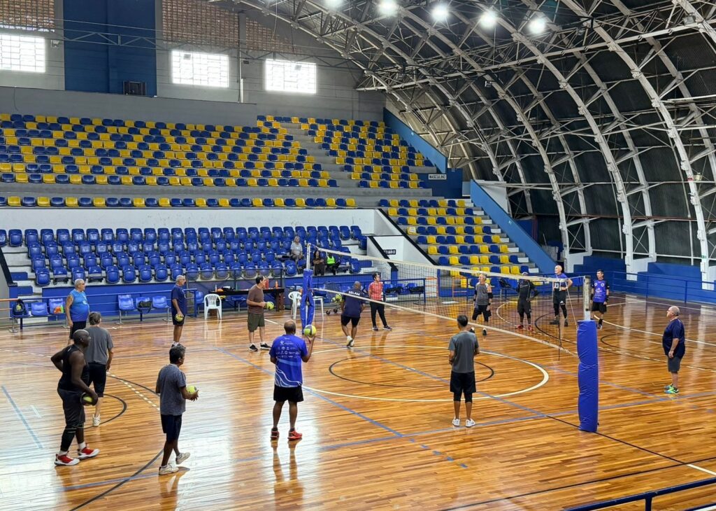 A imagem mostra um grupo de homens jogando voleibol em uma quadra interna. O ginásio possui arquibancadas azuis e amarelas, com iluminação artificial. A quadra é de madeira polida, com as marcações típicas de um jogo de voleibol. Os jogadores estão vestidos com roupas esportivas e alguns seguram bolas de voleibol. Há uma rede montada no centro da quadra, dividindo-a em duas metades. A atmosfera geral sugere uma partida de lazer ou treino.