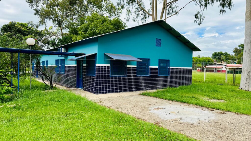 A imagem mostra um edifício de cor azul-petróleo com uma base de azulejos pretos e brancos. O telhado é de metal e inclinado. Há várias janelas com persianas azuis e toldos sobre algumas delas. Uma passarela azul com um poste de iluminação branco com uma lâmpada esférica se estende pela lateral do edifício. A grama verde e exuberante cobre o chão ao redor do edifício, e um caminho de concreto leva até ele. Ao fundo, ao longe, é possível ver um campo com cercas e outros edifícios. O céu está parcialmente nublado, com nuvens brancas e azuis. Grandes árvores com folhagem verde e troncos claros também são visíveis na paisagem.