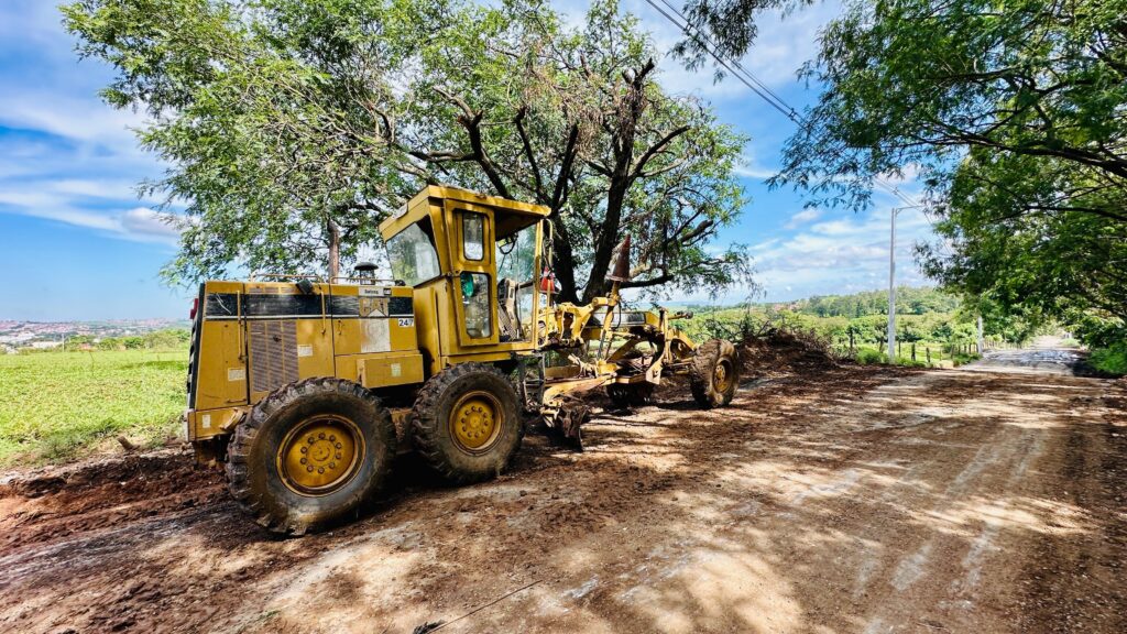 A imagem mostra um *motoniveladora amarela* em um *terreno de terra batida, possivelmente em **obras de construção ou manutenção de estradas. A máquina, que parece ser da marca **CAT, está posicionada sob a sombra de uma **árvore frondosa*, com o sol brilhando intensamente, criando um contraste entre a luz e a sombra. Ao fundo, observa-se uma *paisagem rural, com **colinas verdes, **pastagens* e algumas *construções distantes, sugerindo que o local está em uma área de transição entre o campo e a cidade. O céu está **azul, com algumas **nuvens brancas, indicando um dia ensolarado. A estrada à frente está **esburacada e lamacenta, com montes de terra e detritos ao lado, reforçando a ideia de trabalho em andamento. Há também **postes de iluminação pública* e *fios elétricos* visíveis, indicando a proximidade de uma área habitada.