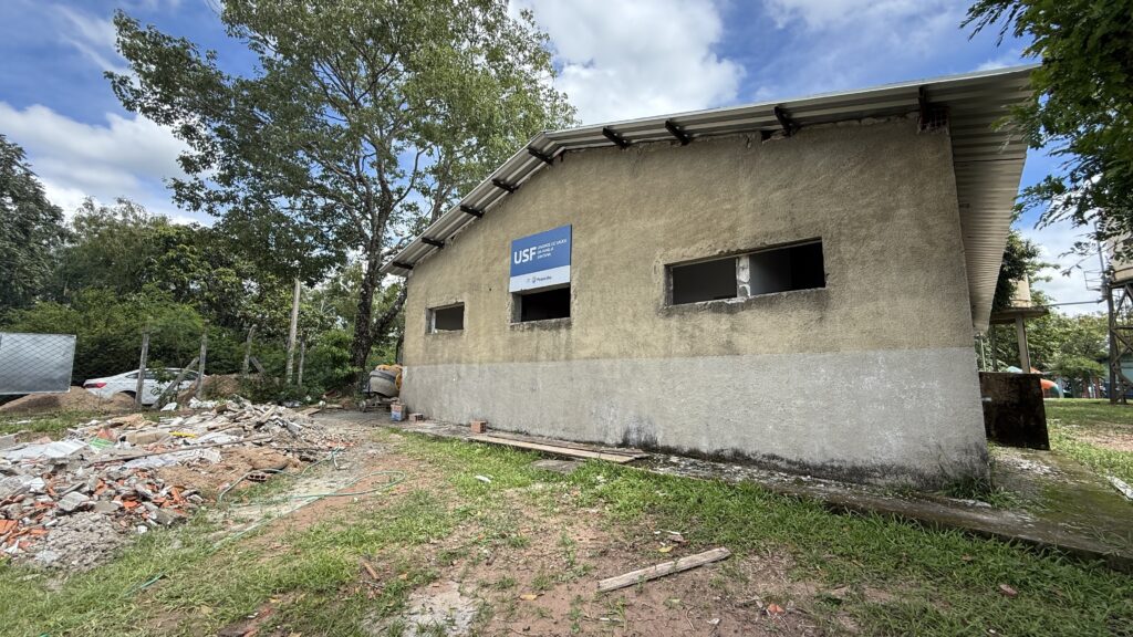 A imagem mostra um prédio de um único andar com uma placa azul e branca que diz "USF UNIDADE DE SAUDE DA FAMILIA". O prédio parece estar em construção ou reforma, com janelas vazias e uma pilha de escombros na frente. Há uma árvore grande à esquerda e vegetação ao redor. O céu está nublado.