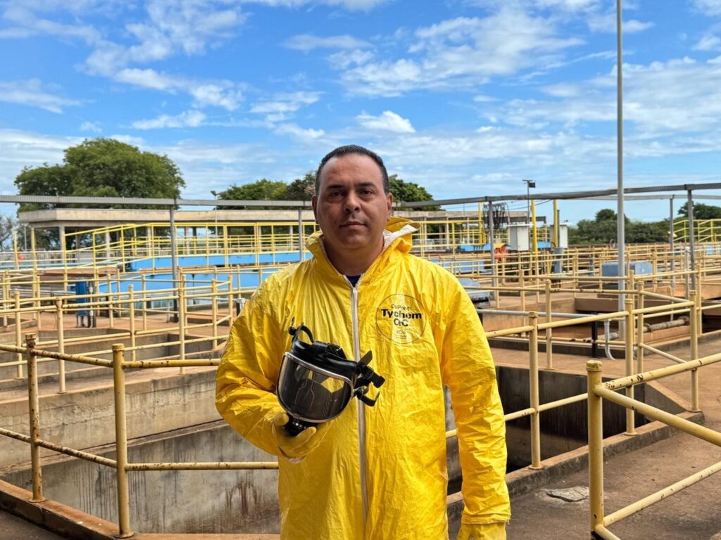 Um homem de meia-idade, vestindo um macacão amarelo brilhante da DuPont Tychem QC, está parado em frente a uma estação de tratamento de água. Ele segura uma máscara preta e transparente na mão direita. O macacão parece ser de corpo inteiro, com capuz e luvas integrados. A máscara, que ele segura, cobre o rosto e tem lentes transparentes e um respirador. O fundo da imagem mostra a infraestrutura de uma estação de tratamento de água, com tanques de concreto, tubulações e passarelas metálicas amarelas. O céu está azul com algumas nuvens brancas, sugerindo um dia ensolarado. A imagem transmite uma sensação de profissionalismo e segurança, com o homem preparado para trabalhar em um ambiente que pode exigir proteção.