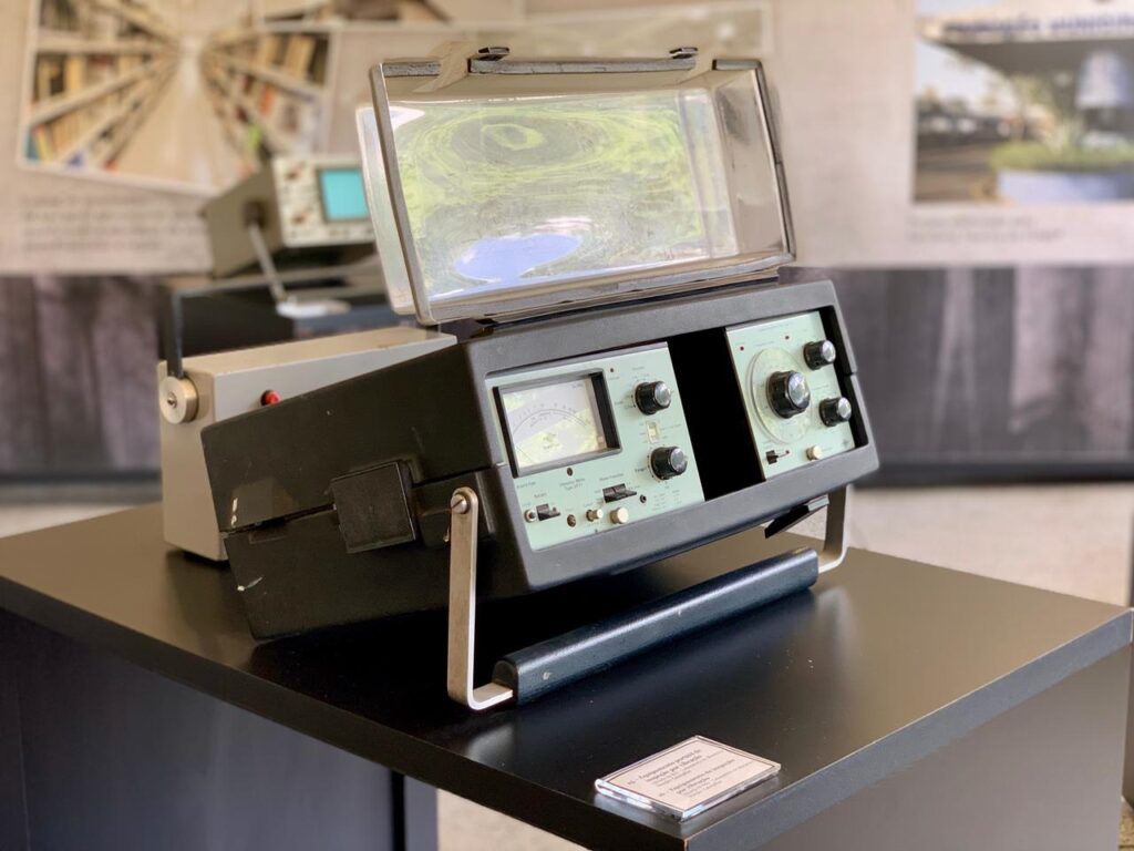 A imagem mostra um equipamento eletrônico antigo, possivelmente um contador Geiger ou um dispositivo de medição similar, em exposição sobre um pedestal preto. O aparelho principal é preto, com um painel frontal verde claro, onde se destacam um medidor analógico, vários botões e um dial circular com botões menores ao redor. Uma tampa transparente, levemente inclinada, cobre a parte superior do painel. À esquerda do equipamento principal, há um outro aparelho cinza, com um botão vermelho visível. Ao fundo, um display de osciloscópio e um painel com imagens desfocadas, possivelmente de uma biblioteca e uma estrutura arquitetônica, criam um ambiente de museu ou exposição. Uma pequena placa informativa está posicionada na frente do pedestal.