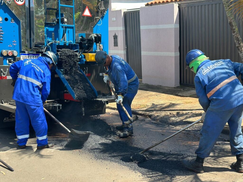 A imagem mostra três trabalhadores da construção civil realizando a pavimentação de uma rua residencial. Eles estão usando uniformes azuis e capacetes de segurança. À esquerda, há uma máquina grande e azul para pavimentação, que está despejando asfalto quente na via. Um dos trabalhadores usa uma pá para espalhar o asfalto próximo à máquina, enquanto outro, no centro, utiliza uma ferramenta longa para nivelar o material. O terceiro trabalhador, à direita, está usando um ancinho para alisar o asfalto fresco. Ao fundo, observa-se um muro rosa com um portão metálico escuro e algumas plantas. O dia está ensolarado, com sombras visíveis no chão. No uniforme de um dos trabalhadores está escrito "Prefeitura do Município de Piracicaba" e em outro uniforme aparece a marca "Molise Serviços & Construções Ltda". A cena retrata o trabalho manual e o uso de equipamentos para manutenção urbana.