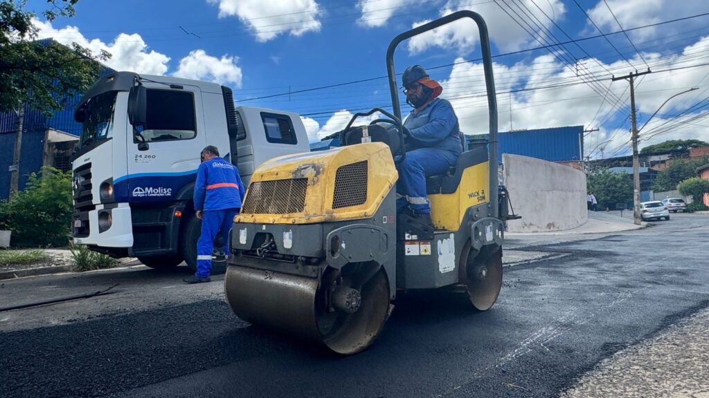 A imagem mostra uma cena de pavimentação de estrada durante o dia, com céu azul claro e algumas nuvens brancas. No centro da imagem, há um rolo compactador vibratório amarelo e preto da marca Wacker Neuson, que está sobre uma camada recém-aplicada de asfalto escuro. Um operador, vestido com uniforme azul, capacete de segurança branco e proteção laranja no pescoço, está sentado no rolo, conduzindo a máquina. À esquerda, há um caminhão branco da marca Mercedes-Benz com a inscrição "Molise Serviços e Construções Ltda." na porta, indicando que a empresa responsável pela obra provavelmente é a Molise. Perto do caminhão, outro trabalhador com uniforme similar está em pé, observando a operação. O ambiente parece ser uma rua residencial ou industrial, com muros de concreto e vegetação ao fundo. Linhas de energia elétrica cruzam o céu, e ao fundo da via há um carro branco estacionado. A iluminação natural destaca os detalhes do asfalto, da maquinaria e das roupas dos trabalhadores, transmitindo uma sensação de um dia claro e produtivo de trabalho.