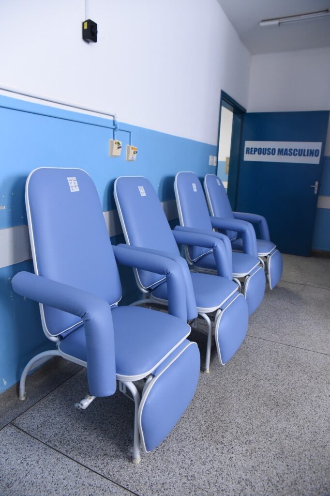 A imagem mostra uma área interna de uma unidade de saúde, provavelmente um hospital ou clínica, dedicada ao repouso masculino, conforme indica a placa na porta ao fundo com o texto "REPOUSO MASCULINO". Na parede à esquerda, há quatro poltronas reclináveis azuis alinhadas em sequência, com estrutura metálica branca. As poltronas possuem apoio para os braços e para as pernas, sugerindo conforto para pacientes que aguardam ou se recuperam. A parede está pintada em duas cores: a parte superior é branca e a inferior azul, combinando com as poltronas e a porta. O ambiente está bem iluminado, com piso claro e limpo, transmitindo uma sensação de organização e higiene. Não há pessoas na imagem, apenas o mobiliário e a ambientação do espaço.