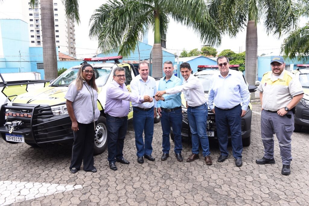 A imagem mostra um grupo de sete pessoas posando para uma foto em um ambiente externo, possivelmente em um estacionamento ou pátio. No centro, cinco homens estão alinhados, segurando juntos uma chave grande, simbolizando a entrega ou recebimento de veículos. À esquerda, uma mulher com óculos e camisa cinza observa a cena, e à direita, um homem vestido com uniforme de trânsito, incluindo boné, está ao lado dos veículos. Ao fundo, há dois veículos tipo caminhonete utilitária, pintados em branco e amarelo, equipados com barras de proteção frontais e luzes no teto, provavelmente veículos oficiais de trânsito. Um dos veículos tem a placa "UFK0I19" visível. O entorno inclui algumas palmeiras e construções urbanas ao longe, sob um céu claro e com iluminação natural. A foto transmite um momento formal, possivelmente a entrega oficial dos veículos para uso em atividades de fiscalização ou patrulhamento.