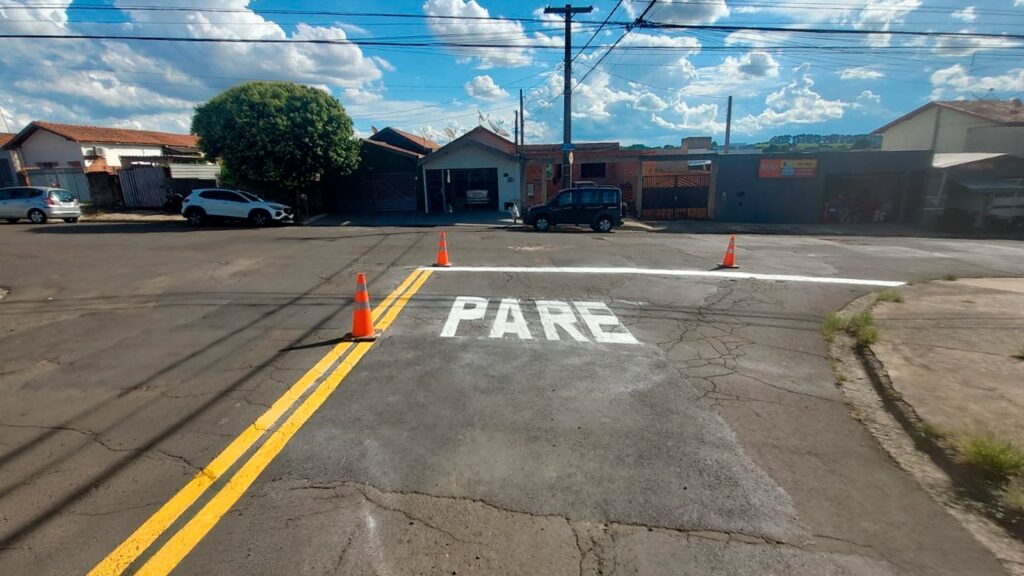 A imagem mostra uma rua residencial durante o dia, com um sinal de trânsito pintado no chão que diz "PARE" em letras brancas grandes e visíveis. A palavra está pintada logo antes de uma linha de parada branca, indicando um ponto onde os veículos devem parar. Ao redor da pintura recém-feita, há cones de trânsito laranja com faixas refletivas, provavelmente para proteger a pintura fresca e alertar os motoristas para não passarem por cima. A rua é asfaltada, com uma linha amarela dupla à esquerda, e ao fundo há alguns carros estacionados e casas típicas de bairro residencial. O céu está parcialmente nublado, e a iluminação indica que é um dia claro e ensolarado. A cena transmite uma sensação de manutenção urbana recente, com atenção à segurança no trânsito local.