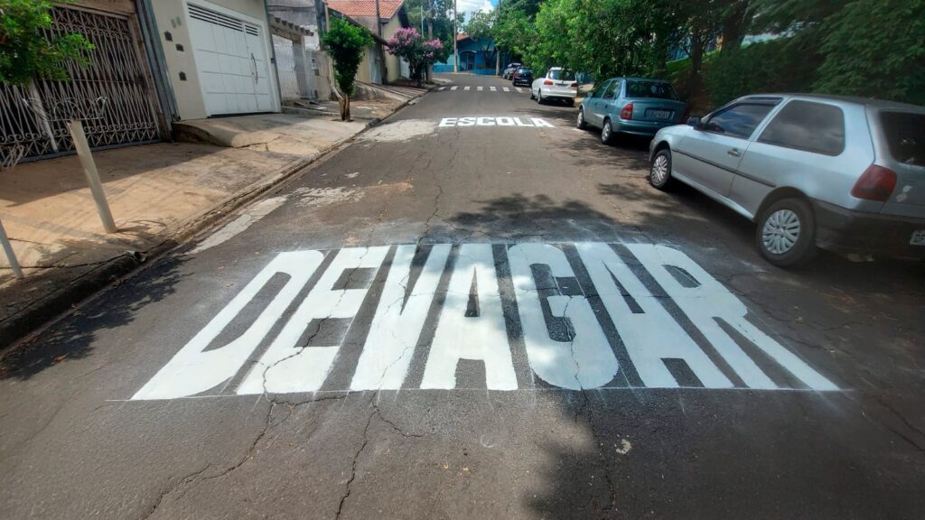 A imagem mostra uma rua residencial tranquila durante o dia, com várias casas e carros estacionados ao longo do lado direito da via. No chão, em letras grandes e brancas, estão pintadas as palavras "DEVAGAR" e "ESCOLA", indicando que é uma área escolar onde os motoristas devem reduzir a velocidade para garantir a segurança. A palavra "DEVAGAR" está mais próxima do observador, enquanto "ESCOLA" aparece um pouco mais adiante, perto de uma faixa de pedestres. A rua é cercada por árvores e casas com muros e portões. O céu está claro, e a iluminação natural destaca o contraste entre a pintura branca no asfalto escuro. A imagem transmite uma sensação de calma e alerta para a importância de dirigir com cuidado em áreas próximas a escolas.