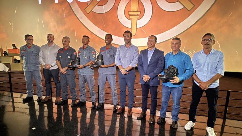 A foto mostra nove homens alinhados no palco durante uma cerimônia formal. Entre eles, alguns vestem uniformes cinza-azulados, possivelmente de bombeiros ou forças de segurança, enquanto outros estão com roupas civis, como camisa social e blazer. Três dos homens exibem botas táticas pretas, sugerindo que o evento está relacionado à entrega ou apresentação desse equipamento. Ao fundo, um grande telão exibe um emblema circular com uma espada e elementos cruzados sobre um fundo que lembra fogo, reforçando a temática de segurança ou combate a incêndios. O palco é escuro e reflete as imagens dos presentes, com iluminação clara voltada para eles. À esquerda, há um suporte com mais botas expostas. A imagem transmite um momento oficial de cooperação ou doação de equipamentos para as forças de segurança ou corpo de bombeiros.