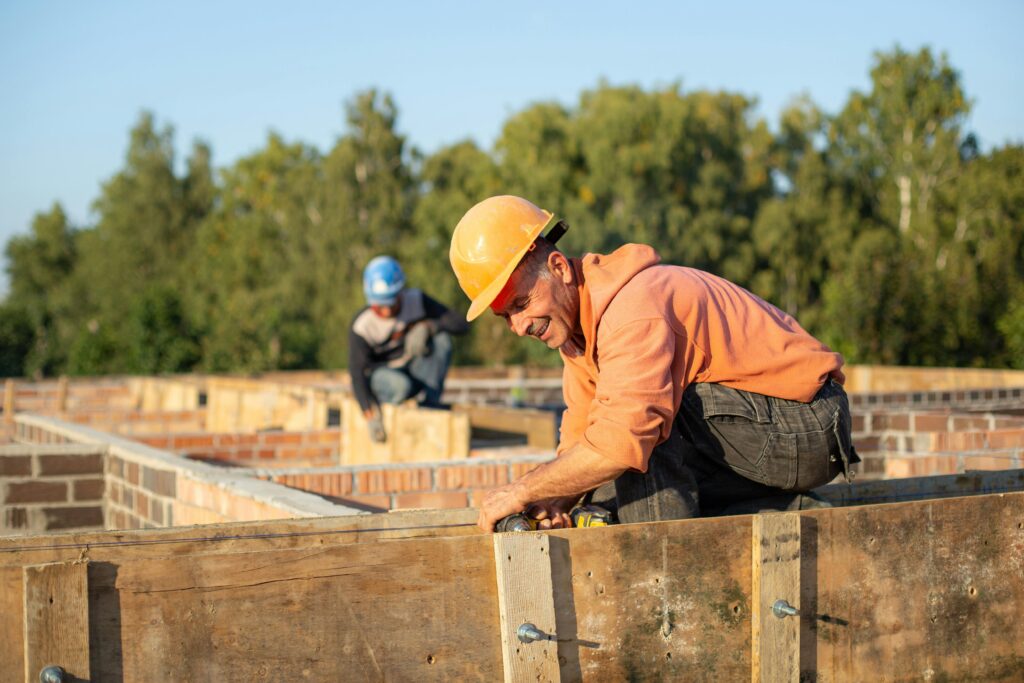 A imagem mostra um canteiro de obras ao ar livre, onde dois trabalhadores estão envolvidos na construção de uma estrutura, provavelmente uma fundação. Em primeiro plano, um homem mais velho, usando capacete e moletom laranja, está ajoelhado sobre uma estrutura de madeira (forma de concreto) e utilizando uma ferramenta elétrica, possivelmente uma furadeira ou parafusadeira. Ele parece concentrado e está ligeiramente sorrindo. Ao fundo, desfocado, há outro trabalhador com capacete azul, agachado, também focado no trabalho. O cenário ao redor inclui paredes de alvenaria e uma linha de árvores verdes sob um céu azul claro, indicando um dia ensolarado. A imagem transmite uma sensação de trabalho em equipe, esforço e progresso na construção.