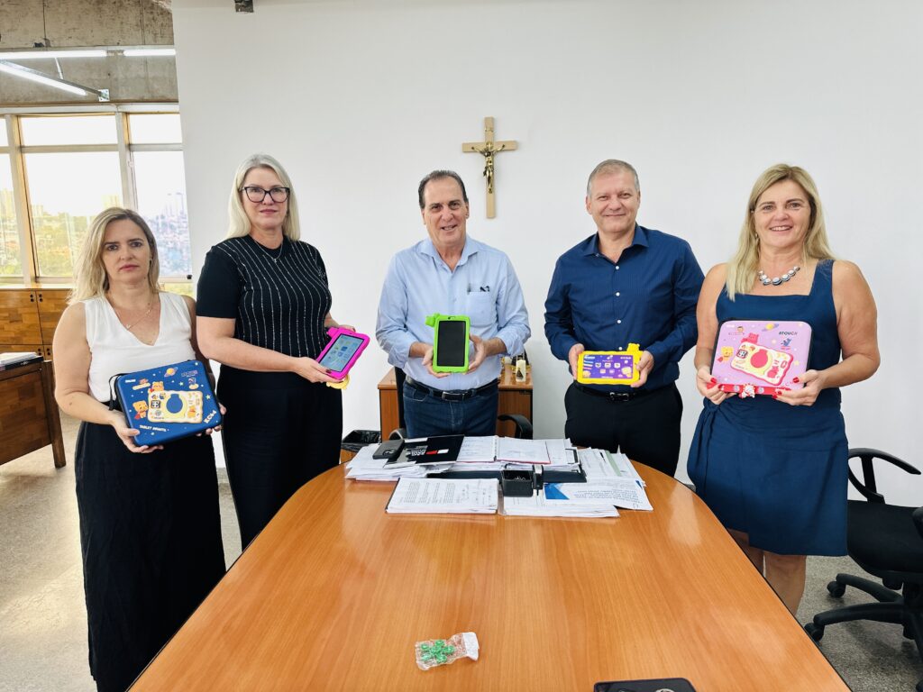 A imagem mostra cinco adultos em um ambiente de escritório, todos posicionados atrás de uma grande mesa oval de madeira polida. Eles estão alinhados e seguram tablets infantis coloridos, cada um com uma capa diferente (azul, rosa, verde e amarelo). As pessoas estão sorrindo, indicando um momento de celebração ou apresentação. Na mesa, há alguns documentos e pastas organizadas. Ao fundo, na parede branca, há um crucifixo pendurado, sugerindo que o local pode ser uma instituição com influência religiosa ou um ambiente formal. À esquerda, uma janela grande permite a entrada de luz natural, mostrando uma vista urbana ao fundo. A cena parece retratar a entrega oficial de tablets infantis, possivelmente para fins educacionais.