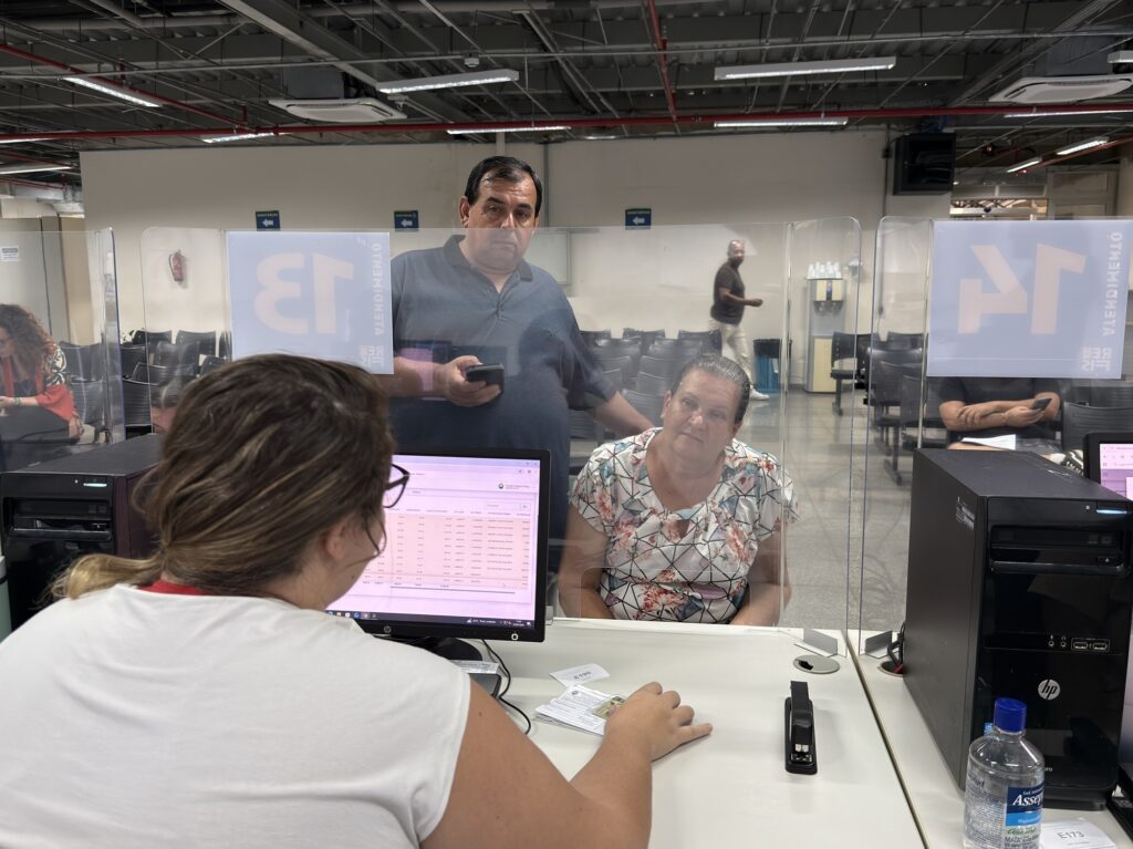 A imagem mostra um ambiente de atendimento ao público, possivelmente em um órgão governamental ou uma repartição pública no Brasil, dado que o texto está em português. No primeiro plano, uma mulher de cabelos castanhos, vestindo uma camisa branca, está de costas para a câmera, interagindo com um casal do outro lado de um balcão. O casal parece estar solicitando algum serviço ou informação; o homem segura um celular, enquanto a mulher está sentada. O balcão é equipado com computadores HP, teclados e monitores, além de um acrílico transparente que funciona como proteção entre os funcionários e os clientes. Há também objetos como um grampeador e uma garrafa de água sobre a mesa. No fundo, é possível ver cadeiras alinhadas, indicando uma área de espera para os usuários. Na parte superior do acrílico, há placas com os números "13" e "14" e a palavra "ATENDIMENTO", indicando os guichês ou postos de atendimento. Também se vê o nome "Asseptgel", sugerindo a presença de álcool em gel para higiene, e palavras como "SANITÁRIOS", provavelmente indicando a direção dos banheiros. O ambiente é bem iluminado, com luz branca fluorescente, e transmite uma atmosfera típica de serviços públicos, organizada e funcional.
