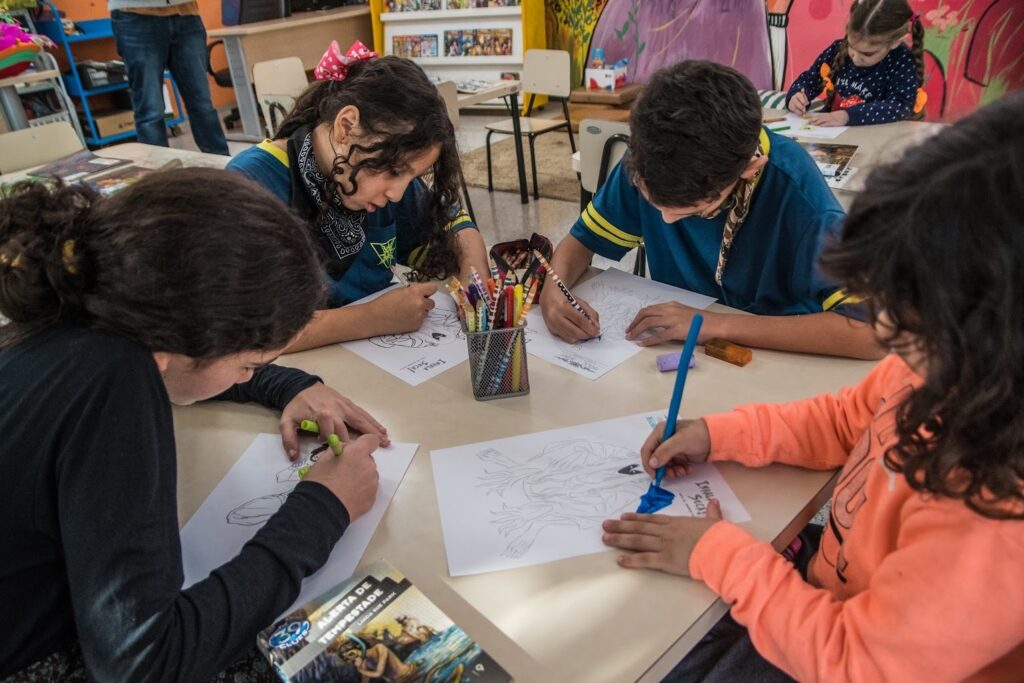A imagem mostra quatro crianças sentadas ao redor de uma mesa retangular clara, concentradas em desenhar e colorir em folhas de papel. No centro da mesa há um recipiente metálico cheio de lápis de cor. Cada criança está focada na sua atividade artística, com desenhos à sua frente, incluindo um que tem a inscrição "IRALA SECA!". As crianças vestem roupas coloridas: uma está de camiseta azul com detalhes amarelos, outra de suéter laranja, uma terceira com uma blusa preta e outra com camiseta azul clara e lenço na cabeça. Ao fundo, é possível ver uma estante com livros e uma parede com decoração colorida, sugerindo que o ambiente é uma sala de aula ou biblioteca. Na mesa em primeiro plano, há um livro intitulado "THE 39 CLUES - ALERTA DE TEMPESTADE" da autora Linda Sue Park, indicando que a atividade pode estar relacionada à leitura ou a um projeto educacional. A luz natural e o ambiente acolhedor transmitem uma atmosfera de aprendizado e criatividade.