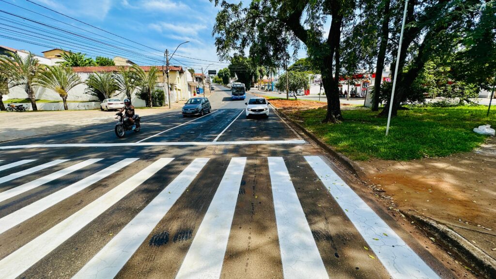 A imagem mostra uma cena urbana durante o dia, com céu azul e algumas nuvens. Em primeiro plano, há uma faixa de pedestres (zebra) bem visível, pintada em branco sobre o asfalto escuro da rua. Na via, circulam alguns veículos: um carro cinza da Volkswagen, um carro branco da Fiat, um ônibus azul com o letreiro "210 COHAB / TPI" e uma motocicleta com um motociclista usando capacete. À esquerda, há casas com telhados vermelhos e muros baixos, enquanto à direita, grandes árvores verdes margeiam a calçada, conferindo sombra e um aspecto mais natural ao ambiente. A rua é relativamente larga e parece ser uma via principal da região. O cenário sugere um bairro residencial ou misto, possivelmente no Brasil, dado o estilo dos veículos e a linguagem do letreiro do ônibus. A composição da foto é ampla, com boa iluminação natural, destacando o contraste entre o asfalto e as faixas brancas da faixa de pedestres, além da vegetação e as construções ao redor. A imagem transmite uma sensação de cotidiano urbano tranquilo.