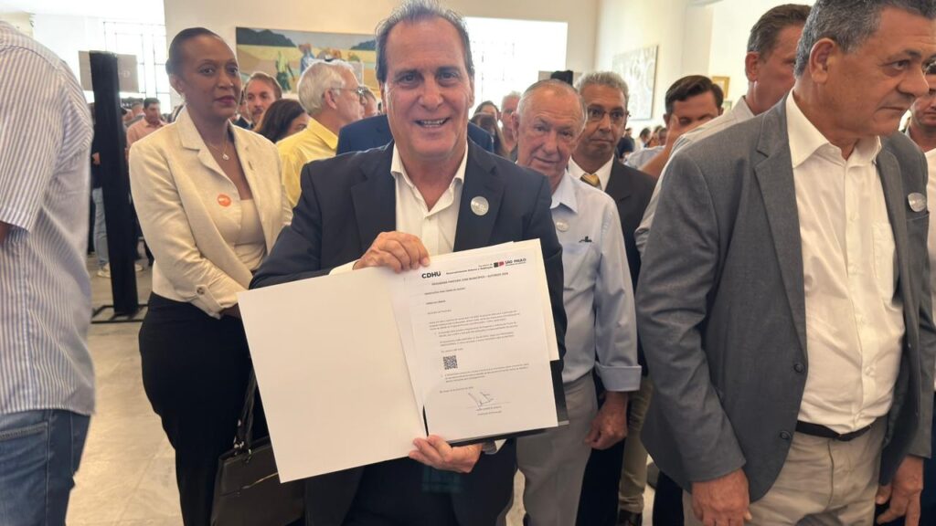 A imagem mostra um grupo de pessoas em um ambiente interno, possivelmente durante uma cerimônia oficial ou evento público. No centro da foto, um homem de meia-idade, vestido com blazer azul escuro e camisa branca, sorri amplamente enquanto segura um documento aberto, que apresenta o logo da CDHU (Companhia de Desenvolvimento Habitacional e Urbano do Estado de São Paulo) e o título "Programa Parceria com Municípios - Autoriza 2024". O documento parece ser um termo de ciência relacionado à adesão do município ao programa. Ao redor dele, outras pessoas também vestem roupas formais, como blazers e camisas, e parecem estar participando do evento. Ao fundo, uma pintura de cena rural decora a parede, e mais pessoas são vistas ao longe, sugerindo que o evento é de certa importância institucional. O ambiente está bem iluminado, transmitindo um clima profissional e de celebração, indicando que o momento registrado é de anúncio ou assinatura de um acordo ou projeto oficial entre o município e a CDHU.