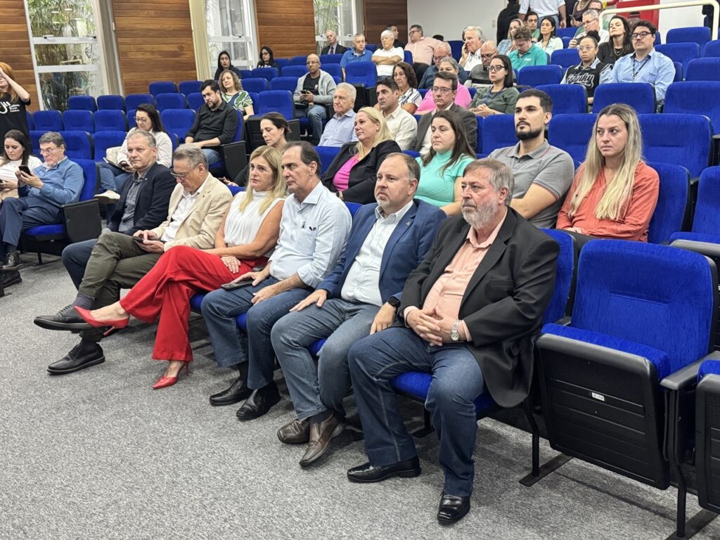 A imagem mostra um grupo de pessoas sentadas em um auditório ou sala de conferências, com várias fileiras de cadeiras acolchoadas azuis. No foco principal, há cerca de sete pessoas na primeira fila, todas olhando para a esquerda, possivelmente assistindo a uma apresentação ou palestra. Entre elas, uma mulher com calça vermelha vibrante e sapatos vermelhos se destaca. Os demais estão vestidos de forma formal ou casual elegante, com blazers e camisas. Atrás delas, outras pessoas também estão sentadas, algumas olhando para frente, outras para seus celulares. O ambiente é iluminado artificialmente, com paredes laterais que têm painéis de madeira e janelas. O chão é revestido com carpete cinza. A atmosfera parece ser de concentração e atenção, típica de um evento profissional ou acadêmico.