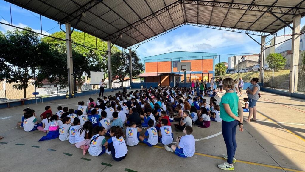 A imagem mostra um grande grupo de estudantes sentados no chão de uma quadra esportiva coberta, provavelmente em uma escola pública. Eles estão organizados em fileiras e vestidos com camisetas brancas que têm o logo colorido da "Educação" e a identificação "Prefeitura de Piracicaba". À frente do grupo, um homem está em pé, aparentemente fazendo uma apresentação ou palestra, com um telão de projeção ao seu lado. Ao redor da quadra, alguns adultos, possivelmente professores ou monitores, acompanham a atividade. O ambiente é iluminado pela luz natural do dia, e ao fundo é possível ver uma construção de tijolos, árvores e algumas casas em uma área residencial. A cena sugere uma atividade educacional coletiva, como uma assembleia, palestra ou evento escolar realizado ao ar livre, mas protegido pelo telhado da quadra.